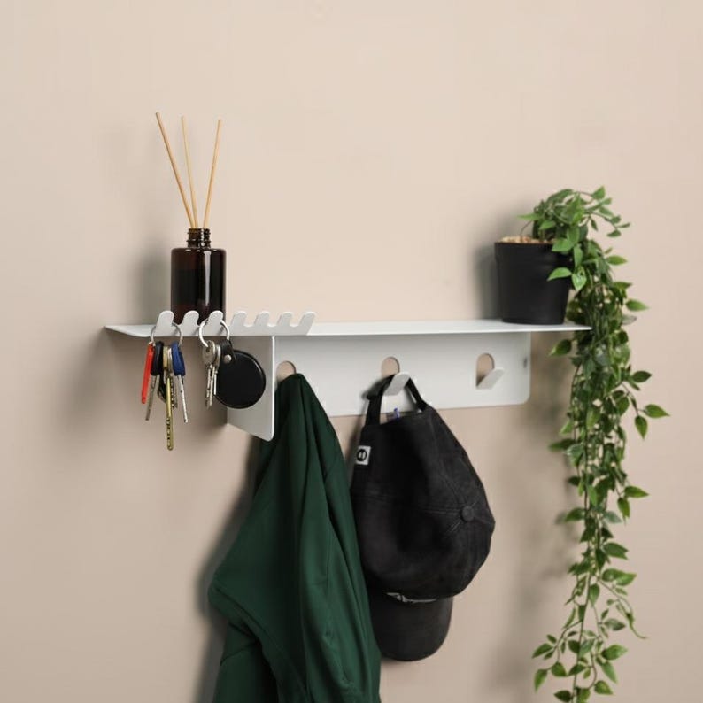 Pu&ograve; includere: Una mensola da parete in metallo bianco con una superficie piana, una fila di ganci integrati per appendere oggetti e una serie di fessure sul lato sinistro. La mensola sostiene un flacone in vetro scuro con bastoncini, un indumento verde e un cappellino da baseball nero.