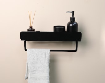 May include: A matte black metal wall-mounted shelf featuring a flat top surface and an integrated towel bar underneath. The shelf holds a small amber glass reed diffuser, a black cup, and a black soap dispenser. A white towel hangs on the bar.
