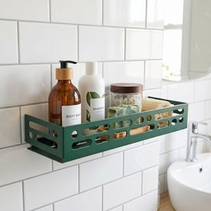 May include: A dark green rectangular metal shower caddy with cut-out slots, holding a brown bottle labeled BODY WASH, a white bottle labeled SHAMPOO, a clear jar with a wooden lid, a bar of soap, and a natural fiber sponge.