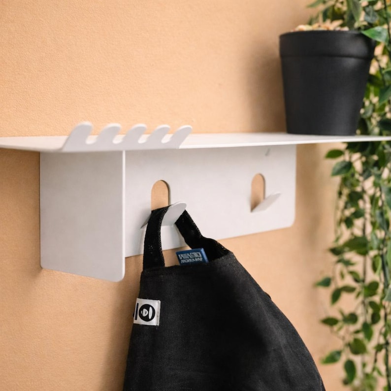 Puede incluir: Un estante de pared de metal blanco con una superficie superior plana con un borde ondulado y dos ganchos integrados debajo. Un delantal de tela negra con una peque&ntilde;a etiqueta rectangular blanca cuelga del gancho izquierdo del estante.