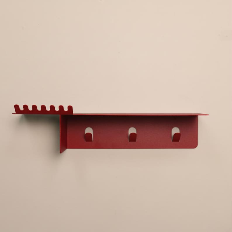 Puede incluir: Un organizador de pared de metal rojo mate que cuenta con un estante plano, una secci&oacute;n lateral con seis peque&ntilde;as muescas para colgar objetos y un panel trasero vertical equipado con tres ganchos integrados para el almacenamiento de art&iacute;culos.