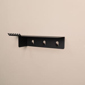 Pu&ograve; includere: Una mensola da parete nero opaco caratterizzata da una superficie superiore piatta con una serie di sei piccole tacche lungo il bordo sinistro e tre ganci integrati che si estendono verso il basso dal pannello orizzontale principale.