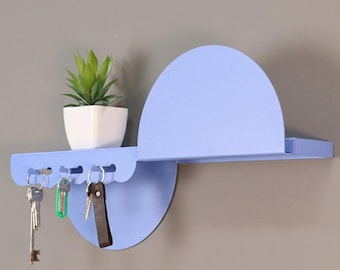 Puede incluir: Un estante de pared de metal de color morado claro con un panel trasero semicircular y una sección inferior con cinco ganchos integrados. Tres llaves de metal cuelgan de los ganchos y una pequeña maceta cuadrada blanca con una suculenta verde descansa encima.
