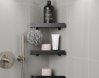 May include: A black corner shower shelf unit with three shelves. The top shelf holds a black shampoo bottle. The middle shelf has a loofah and a tube of cream. The bottom shelf has a jar of cream. The shower wall is gray.