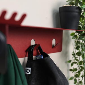 Puede incluir: Un estante de pared de metal rojo mate con una repisa superior plana de borde ondulado y varios ganchos integrados. El estante est&aacute; montado horizontalmente y sostiene una gorra de b&eacute;isbol negra en uno de sus ganchos de metal plateado.