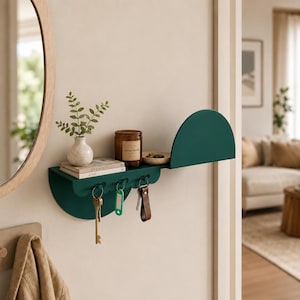 May include: A dark green wall-mounted shelf featuring a semi-circular side panel and three integrated hooks. The shelf holds a small white vase with greenery, a stack of books, a brown candle jar labeled Sandalwood, and a small wooden dish.