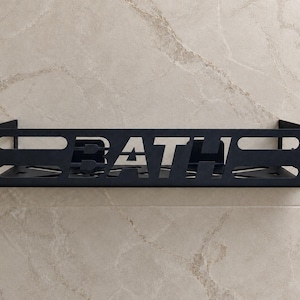 May include: A matte black metal shower caddy featuring a rectangular frame with horizontal cutouts and the word BATH cut out in the centre of the front panel. The shelf has a flat base and side walls to hold items securely in place.