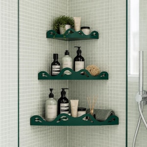May include: Three dark green corner shelves with a scalloped front edge, mounted in a tiled shower. The shelves hold various skincare bottles, a small potted plant, a textured sponge, a tube of cream, and a folded grey washcloth.