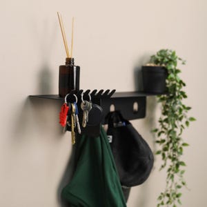 Pu&ograve; includere: Una mensola da parete in metallo nero con una superficie superiore per piccoli oggetti, una fila di ganci per chiavi e ganci aggiuntivi per appendere accessori. La mensola sostiene una bottiglia di vetro marrone con bastoncini e una pianta.