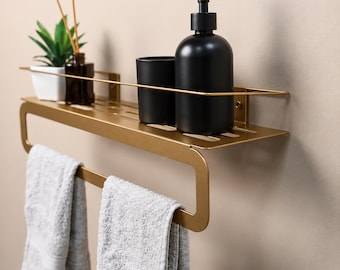 May include: A gold-toned metal wall-mounted bathroom shelf featuring a top storage rack with a protective rail and an integrated towel bar below. Two light gray towels hang from the bar, while a black soap dispenser and cup sit on top.