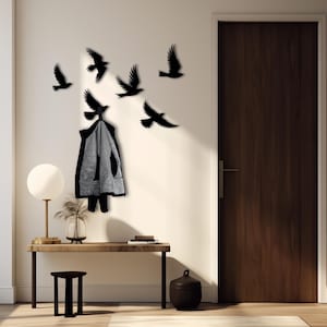 Op de afbeelding: Een zwarte metalen wandbevestigde kapstok met een silhouet van vliegende vogels. Een zwart jack hangt aan de kapstok. De kapstok is bevestigd aan een witte muur met een houten deur aan de rechterkant.