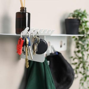 Pu&ograve; includere: Una mensola da parete in metallo bianco dotata di una superficie superiore per riporre oggetti e una serie di ganci integrati lungo il bordo anteriore. La mensola sostiene un flacone in vetro marrone scuro con bastoncini e varie chiavi.