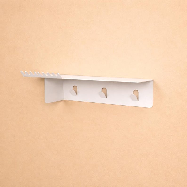 Puede incluir: Un estante de pared blanco con una superficie superior plana y un panel trasero vertical. El estante incluye una fila de peque&ntilde;as muescas en el lado izquierdo y tres ganchos integrados espaciados uniformemente en el panel vertical.