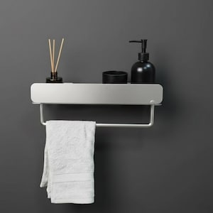 Peut inclure: Étagère de salle de bain moderne argentée avec porte-serviettes. Elle contient un distributeur de savon noir, un petit récipient noir et un flacon diffuseur avec des bâtonnets. Une serviette blanche pliée est suspendue. Fond gris uni.