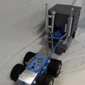 May include: A detailed model of a semi-truck. The truck features a blue chassis, black tires, and a dark gray cab. Chrome exhaust pipes and other metallic details add to the realism of the miniature vehicle. The model is designed to replicate a heavy-duty truck.