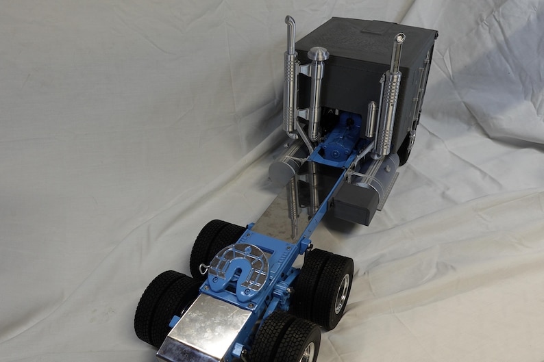 May include: A detailed model of a semi-truck. The truck features a blue chassis, black tires, and a dark gray cab. Chrome exhaust pipes and other metallic details add to the realism of the miniature vehicle. The model is designed to replicate a heavy-duty truck.
