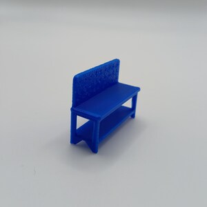 May include: A miniature blue bench featuring a solid seat, a lower shelf, and a backrest with a textured, repeating pattern. The structure is supported by four legs and is rendered in a vibrant, solid blue color with a matte finish throughout.