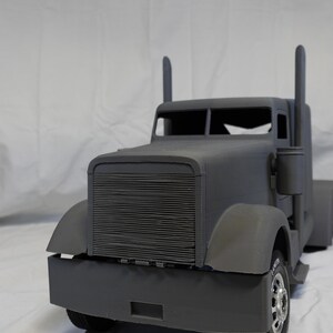 May include: A grey toy articulated lorry with a detailed front grille, mudguards, and exhaust stacks. The lorry has a cab with a windscreen and side mirrors. The wheels are visible, and it appears to be a model or replica.
