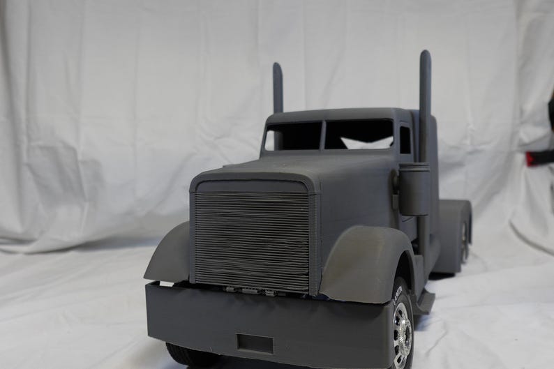 May include: A grey toy articulated lorry with a detailed front grille, mudguards, and exhaust stacks. The lorry has a cab with a windscreen and side mirrors. The wheels are visible, and it appears to be a model or replica.
