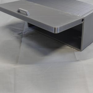 May include: A gray, rectangular storage box with a hinged lid and a small handle. The lid is open, revealing an empty interior. The box is made of a solid material and sits on a white surface.