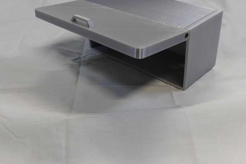 May include: A gray, rectangular storage box with a hinged lid and a small handle. The lid is open, revealing an empty interior. The box is made of a solid material and sits on a white surface.