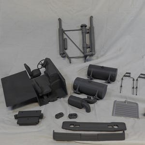 May include: An assortment of gray plastic model truck parts, including a cab with a steering wheel and seat, fuel tanks, and a radiator. The parts are arranged on a white surface.