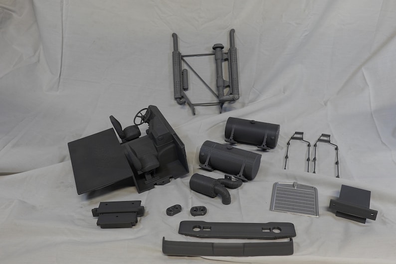 May include: An assortment of gray plastic model truck parts, including a cab with a steering wheel and seat, fuel tanks, and a radiator. The parts are arranged on a white surface.