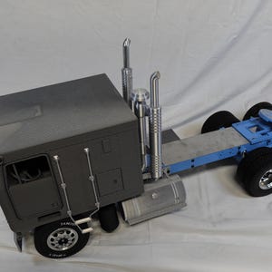 May include: A gray semi-truck model with a blue trailer frame and black tires. The truck has two chrome exhaust stacks and a fuel tank. The wheels have silver rims. The model is on a white surface.