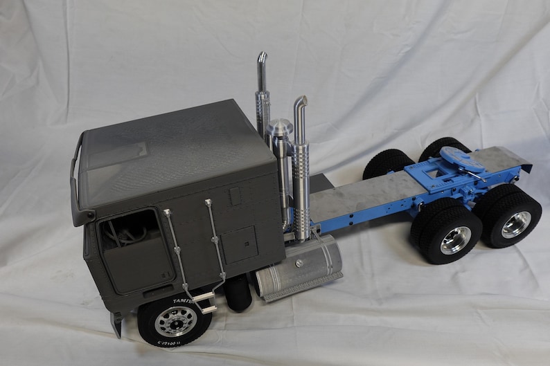 May include: A gray semi-truck model with a blue trailer frame and black tires. The truck has two chrome exhaust stacks and a fuel tank. The wheels have silver rims. The model is on a white surface.