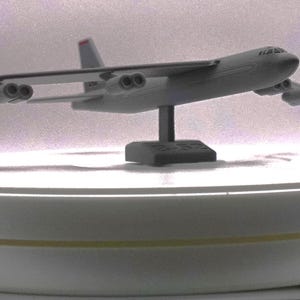 May include: A gray model airplane on a white display stand. The airplane has a long, narrow fuselage and swept wings. It has four engines and a tail fin.