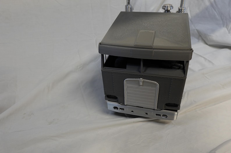 May include: A gray toy semi-truck with a detailed design. The truck features a cab with a roof, windows, and a front grill. The front bumper and other details are visible. The toy is likely made of plastic.