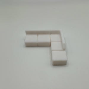May include: A miniature white L-shaped sectional sofa featuring a smooth, matte finish. The piece consists of three connected seat sections and a backrest that extends along two sides, forming a corner configuration with a clean, blocky design.