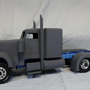 May include: A grey model articulated lorry with a blue chassis. The truck features two exhaust stacks, a cab, and a trailer hitch. The tyres are black with silver rims. The word "TAMIYA" is printed on the tyre.