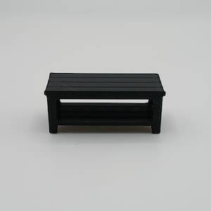 May include: A small, rectangular black bench featuring a slatted top surface and a matching slatted lower shelf. The piece is supported by four sturdy legs, creating a simple, open structure with a uniform matte black finish throughout the design.