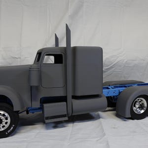 May include: A grey model articulated lorry with a cab, sleeper unit, and trailer hitch. The truck has black tyres with silver rims. The word "TAMIYA" is visible on the tyre. The truck is on a white surface.