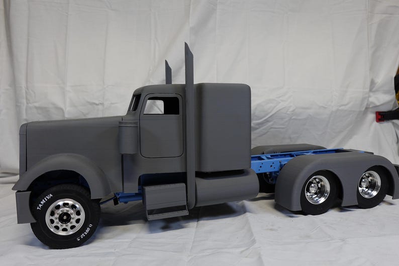 May include: A grey model articulated lorry with a cab, sleeper unit, and trailer hitch. The truck has black tyres with silver rims. The word "TAMIYA" is visible on the tyre. The truck is on a white surface.