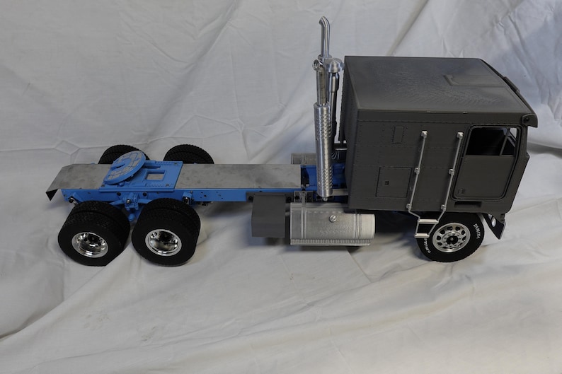 May include: A gray and blue semi-truck model with a detailed design. The truck features a cab, a trailer hitch, and multiple wheels. The model is set against a white background.