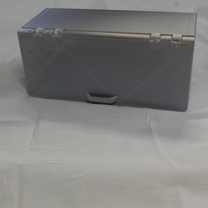 May include: A gray rectangular box with a hinged lid and a small handle on the front. The box has a textured surface and is sitting on a white surface. The box appears to be made of plastic.