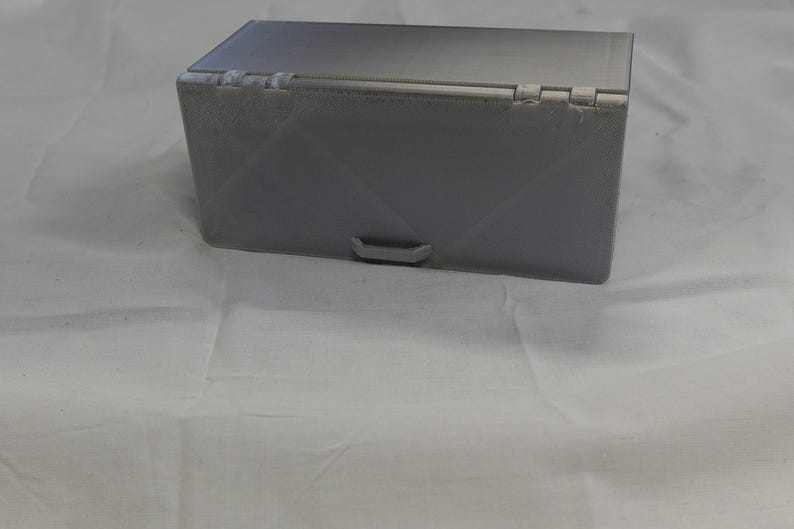 May include: A gray rectangular box with a hinged lid and a small handle on the front. The box has a textured surface and is sitting on a white surface. The box appears to be made of plastic.
