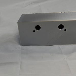 May include: A rectangular, silver-colored metal box with two large circular holes and one small hole on the front. The box has a textured surface and is set against a white background.