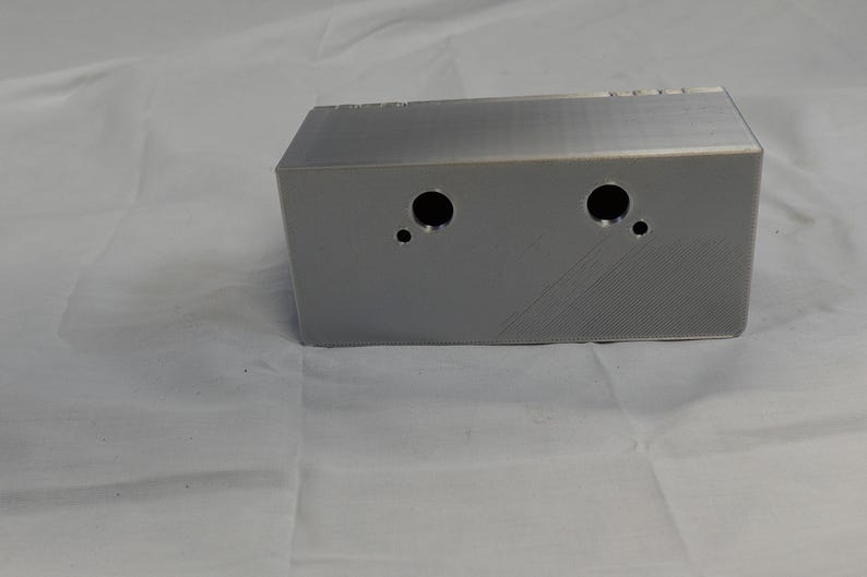 May include: A rectangular, silver-colored metal box with two large circular holes and one small hole on the front. The box has a textured surface and is set against a white background.