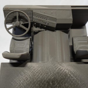 May include: A gray 3D-printed model of a vehicle's interior, featuring a steering wheel, dashboard with gauges, two seats, and a central console. The model is detailed with textured surfaces and appears to be a miniature replica.