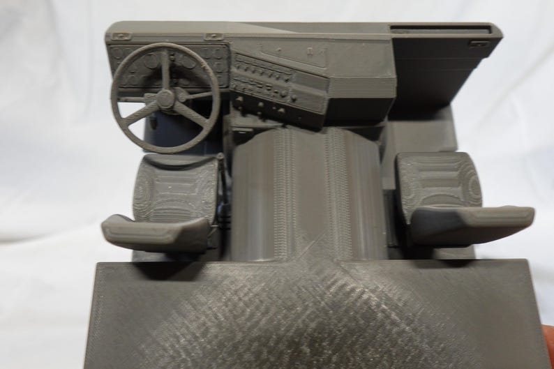 May include: A gray 3D-printed model of a vehicle's interior, featuring a steering wheel, dashboard with gauges, two seats, and a central console. The model is detailed with textured surfaces and appears to be a miniature replica.