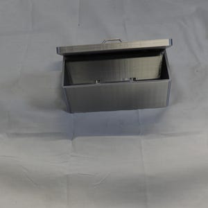 May include: A rectangular, silver-colored box with a hinged lid, open to reveal the interior. The box has a handle on the lid and appears to be made of metal. The box is sitting on a white surface.