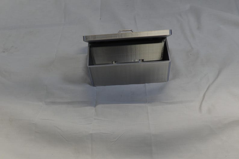 May include: A rectangular, silver-colored box with a hinged lid, open to reveal the interior. The box has a handle on the lid and appears to be made of metal. The box is sitting on a white surface.