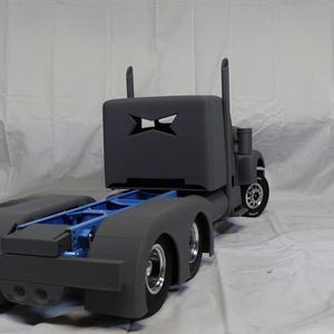 May include: A grey semi-truck model with a blue frame and silver wheels. The truck's cab has a cut-out design. The model is set against a white backdrop, showcasing its details.