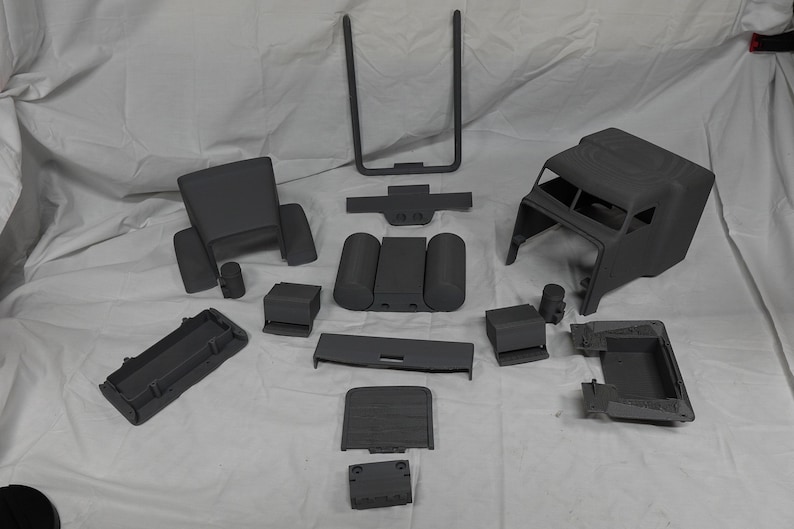 May include: A collection of grey plastic vehicle interior components, including a seat, dashboard parts, and a cab frame, laid out on a white surface. These appear to be model or prototype parts.