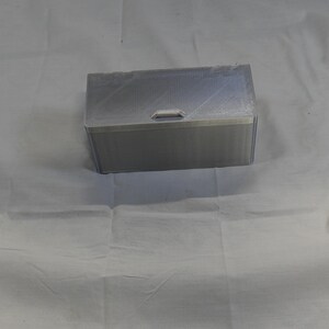 May include: A rectangular, silver-colored box with a clear lid and a small handle. The box appears to be made of a rigid material, possibly plastic or metal. The lid is slightly transparent, and the box is sitting on a white surface.