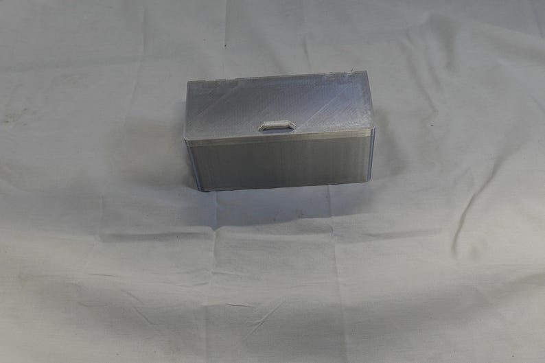May include: A rectangular, silver-colored box with a clear lid and a small handle. The box appears to be made of a rigid material, possibly plastic or metal. The lid is slightly transparent, and the box is sitting on a white surface.