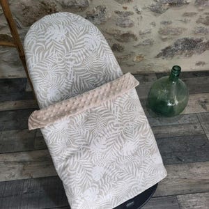 May include: A baby bouncer with a white and beige tropical leaf pattern. The bouncer has a soft, plush, beige border. A green glass bottle sits nearby on a wooden floor.